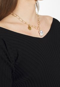 Collar de cadena de oro con eslabones rectangulares, que presenta un colgante de ojo dorado y una gran perla de forma irregular, llevada con una blusa negra off-shoulder.