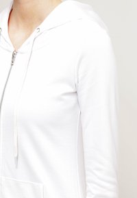 Sudadera blanca con cremallera hecha de algodón suave. Presenta una capucha con cordones, mangas largas y un bolsillo en la parte frontal. Textura suave con un diseño ajustado.