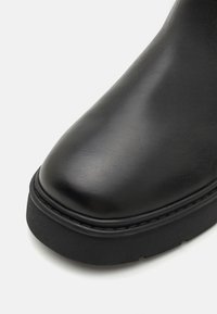 Botte en cuir noir avec un bout arrondi, semelle en caoutchouc noir texturé et des coutures le long du bord. Finition lisse, design minimaliste.