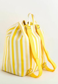Kirkas keltainen ja valkoinen raidallinen reppu, jossa on säädettävät keltaiset hihnat ja ylhäällä oleva kahva, vaaleaa taustaa vasten.