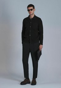 Schwarze Strickjacke mit Knöpfen, einer einzigen Tasche; getragen über einem grauen Oberteil. Schwarze Hose und braune Schuhe, hält eine schwarze Clutch. Glatte Textur, taillierter Schnitt.