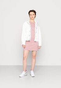 Casaco de algodão branco sobre um vestido às riscas rosa e branco. Modelo a usar ténis brancos, de pé contra um fundo simples.