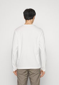 Άτομο που φαίνεται από πίσω με απλό λευκό μακρυμάνικο πουκάμισο και καφέ καρό παντελόνια σε λευκό φόντο.