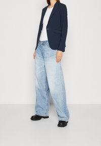 Marinblå blazer över en vit ribbad topp, parat med ljus tvättade vida jeans och svarta skor. Enkel design med minimal detaljering.