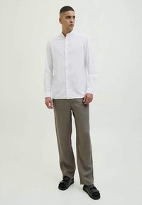 Man standing wearing a white mandarin collar shirt, loose brown trousers, and black sandals against a plain light background.