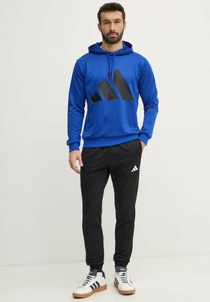 Felpa blu con logo nero, abbinata a pantaloni neri e sneakers bianche. L'outfit presenta un design atletico informale.