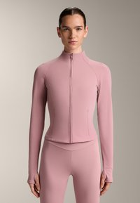 Ljusrosa zip-up jacka med långa ärmar, figursydd design, slät textur och hög krage. Anmärkningsvärda detaljer inkluderar tumhål i muddarna.