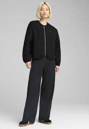 Veste bomber matelassée noire avec fermeture éclair sur le devant, manches à poignet et ourlet arrondi, associée à un pantalon noir ample et des chaussures sombres.