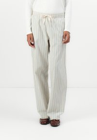 Personne portant un pantalon beige à rayures verticales bleues, un haut à manches longues blanc et des chaussures marron foncé, se tenant devant un fond blanc.