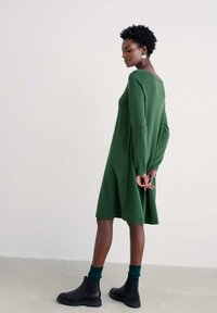 Frau in einem langen, grünen Kleid bis zum Knie, dunklen grünen Socken und schwarzen Stiefeletten, die mit hinten verschränkten Händen dasteht.