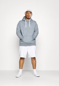 Felpa grigia mélange con cordini bianchi, tasca a marsupio, abbinata a pantaloni corti bianchi e sneakers bianchi, su uno sfondo semplice.