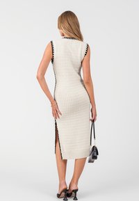 Abito lavorato a maglia crema con rifiniture nere. Design aderente senza maniche con superficie strutturata e fessure laterali. Accessorio borsa nera piccola.
