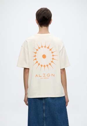 Άτομο με κοντά μαλλιά που φοράει λευκό μπλουζάκι με πορτοκαλί σχέδιο ήλιου και το κείμενο «ALIGN LALA BERLIN» στην πλάτη, συνδυασμένο με μπλε φούστα.