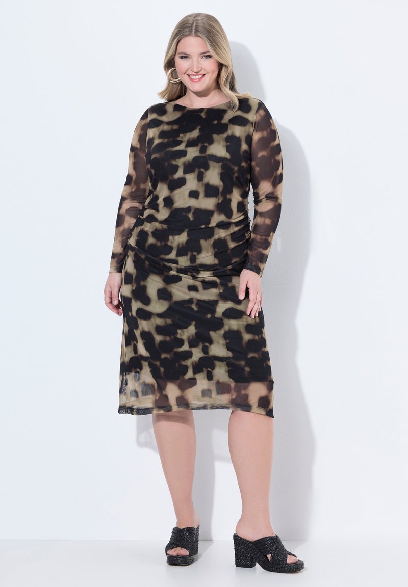 Femme souriante aux cheveux blonds portant une robe imprimé animal noir et olive et des sandales à talons ouvertes noires sur fond blanc.