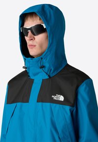 Modrá větrovka s černým panelem přes ramena, nastavitelnou kapucí a bílým logem vpředu. Model má na sobě sluneční brýle.