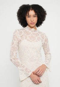 Kiharahiuksinen nainen, jolla on korkea kaulus ja pitkät hihat, kermaan vivahtava pitsipaita, jossa on aaltoilevia kuvioita, seisoo valkoista yksinkertaista taustaa vasten.