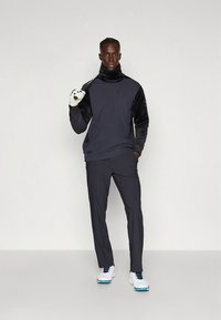 Heren sportoutfit met een donkergrijze top met lange mouwen en contrasterende zwarte accenten, gecombineerd met lichte grijze broek. Witte en blauwe sportschoenen.