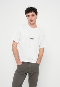 Λευκό βαμβακερό T-shirt με κοντά μανίκια, με μικρό μαύρο λογότυπο στο στήθος. Το σχέδιο είναι σε άνετη εφαρμογή με στρογγυλή λαιμόκοψη.