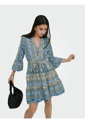 Kobieta ubrana w niebieską sukienkę w wzory z rozszerzanymi rękawami i paskiem, trzymająca czarną marszczoną torebkę, patrząca w dół, z rozpuszczonymi ciemnymi włosami.