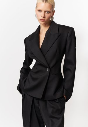 Blondynka ubrana w oversize'owy czarny blazer z podkreślonymi ramionami oraz luźne czarne spodnie, stojąca na białym tle.