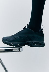 Chaussure de sport noire avec une tige texturée, des accents latéraux et un amorti visible. Design élégant avec un bout arrondi et une semelle en caoutchouc antidérapante.