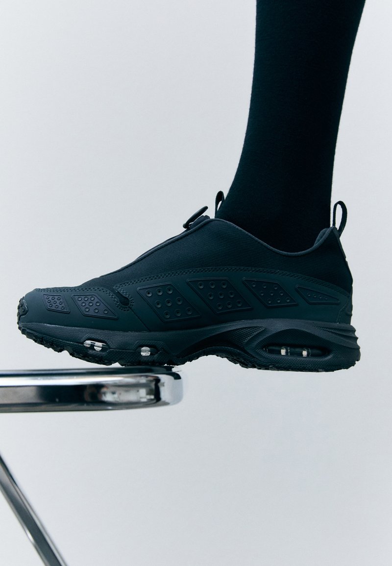 Chaussure de sport noire avec une tige texturée, des accents latéraux et un amorti visible. Design élégant avec un bout arrondi et une semelle en caoutchouc antidérapante.
