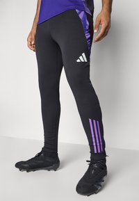 Leggings sportivi neri con accenti laterali viola, caratterizzati da un logo con tre strisce bianche. Realizzati in tessuto elasticizzato per comfort e libertà di movimento.