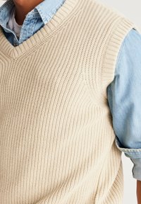 Beżowy sweter bez rękawów z dekoltem w kształcie V i karbowaną teksturą, nałożony na jasnoniebieską koszulę dżinsową z podwiniętymi rękawami.