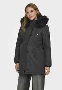 Parka noire avec une capuche en fausse fourrure, fermetures à boutons, taille réglable et poches latérales. Le tissu semble lisse et résistant à l'eau.