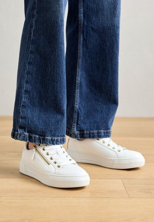 White sneakers with a smooth texture, gold zippers, and eyelets. Paired with flared dark blue denim jeans, slightly cropped at the hem.