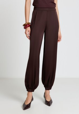 Woman wearing dark brown high-waist tapered pants with elastic cuffs, paired with matching pointed-toe heels and chunky bracelets.
