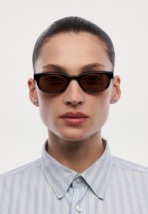 Osoba s uhlazenými vlasy, která nosí úzké černé brýle s rámem a světle modrou pruhovanou košili s knoflíky na neutrálním pozadí.