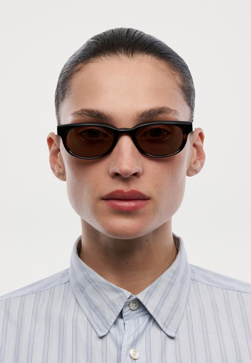 Persona con el cabello peinado hacia atrás, vistiendo gafas de sol estrechas con montura negra y una camisa de botones a rayas azul claro, sobre un fondo neutro.