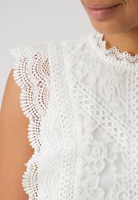 Weißes Spitzenoberteil mit filigranen Blumenmustern, wellenförmigen Kanten und strukturierten Details; verfügt über einen hohen Ausschnitt und ein ärmelloses Design.