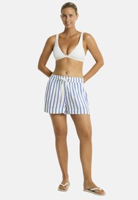 Modelka w białym bikini na górze i niebieskich szortach w paski stoi pewnie, uśmiechając się z rękami na biodrach, na tle jednolitego białego tła.