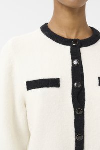 Strikket cardigan i krem med mørkegrå detaljer. Har rund hals, svarte knapper og to svarte lappelommer foran.