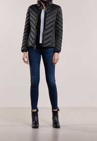 Doudoune noire avec fermeture à glissière sur le devant et col haut, présentant un design matelassé et une silhouette ajustée, assortie à un jean bleu et des bottines noires.