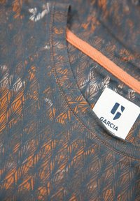 Detailní záběr na tmavě modrou a oranžovou vzorovanou látku s kulatým výstřihem a bílou nálepkou značky Garcia uvnitř límce.