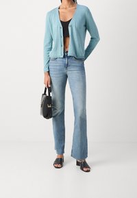 Cardigan bleu clair avec un décolleté en V et une fermeture à boutons, associé à un jean évasé bleu délavé. Un sac à main noir et des sandales noires épaisses complètent la tenue.