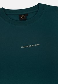 Teal katoenen t-shirt met een ronde hals, met het woord "TOMORROWLAND" in lichtgrijs gedrukt. Minimalistisch ontwerp, zachte textuur.