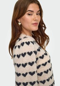 Jersey de punto en suave color crema con patrones de corazones negros, cuello redondo y acabado texturizado. El modelo lleva pendientes en forma de corazón.