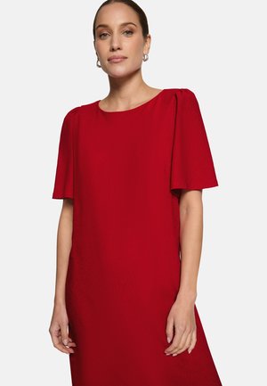 Femme aux cheveux tirés en arrière, portant des boucles d'oreilles argentées en anneau et une robe rouge mi-longue avec des manches courtes légèrement bouffantes.