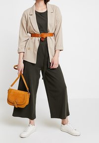 Mujer vestida con blazier beige, pantalones de pierna ancha negros, zapatillas blancas, cinturón marrón, sosteniendo un bolso marrón a juego frente a un fondo blanco.