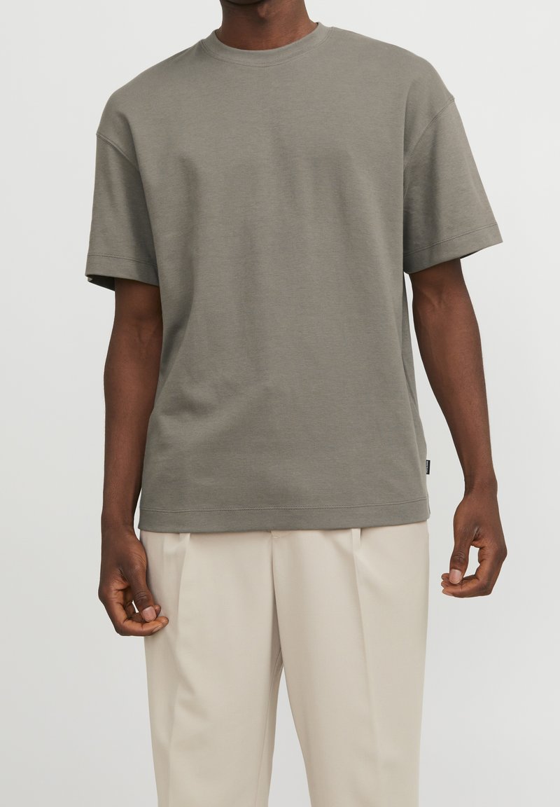 Šedé bavlněné tričko s kulatým výstřihem, spadlými ramenními švy a krátkými rukávy, v kombinaci se světlými béžovými plisovanými kalhotami.