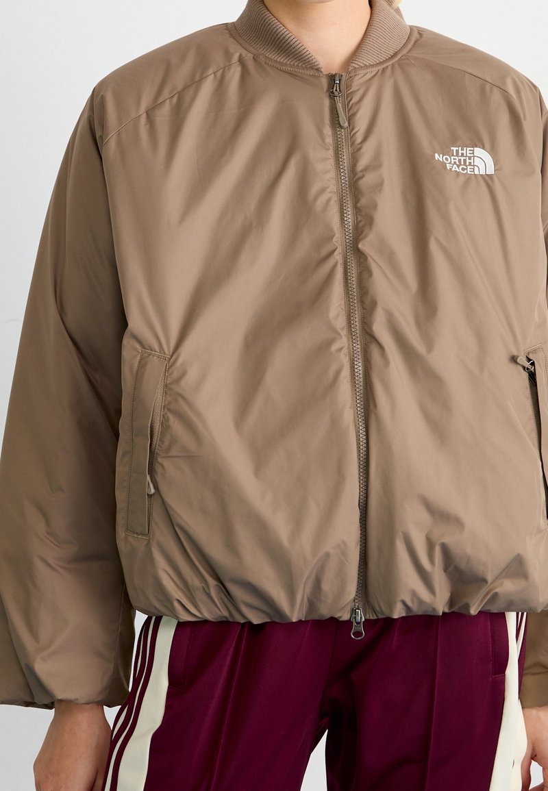 Personne portant une veste à fermeture éclair North Face marron clair associée à un pantalon de survêtement bordeaux avec des bandes blanches.