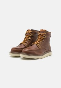 Botas de cuero marrón con cordones amarillos, punta redondeada y costuras en contraste. Presentan un tobillo acolchado y suela de goma beige.