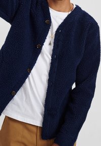 Mörkblå, texturerad fleecejacka med V-ringning, fem knappar och långa ärmar, bärs över en vit T-shirt. Slim fit med en kortad design.