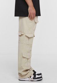 Beige cargo nadrágok, több zsebével és hurkáival, puha anyagból készült. Fekete-fehér tornacipővel viselve. Oldalnézetben látható.