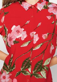 Blusa rossa floreale con motivi di fiori rosa e verdi, dotata di chiusura con bottoni e colletto. Tessuto leggero con una texture liscia.