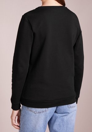 Personne portant un t-shirt noir uni à manches longues et un jean bleu clair, vue de dos sur un fond neutre.